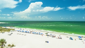 A beach with white sand, clear blue water, and a pier in the distance. - Sandy Shores 504 1 BR Gulf Front (Madeira Beach)