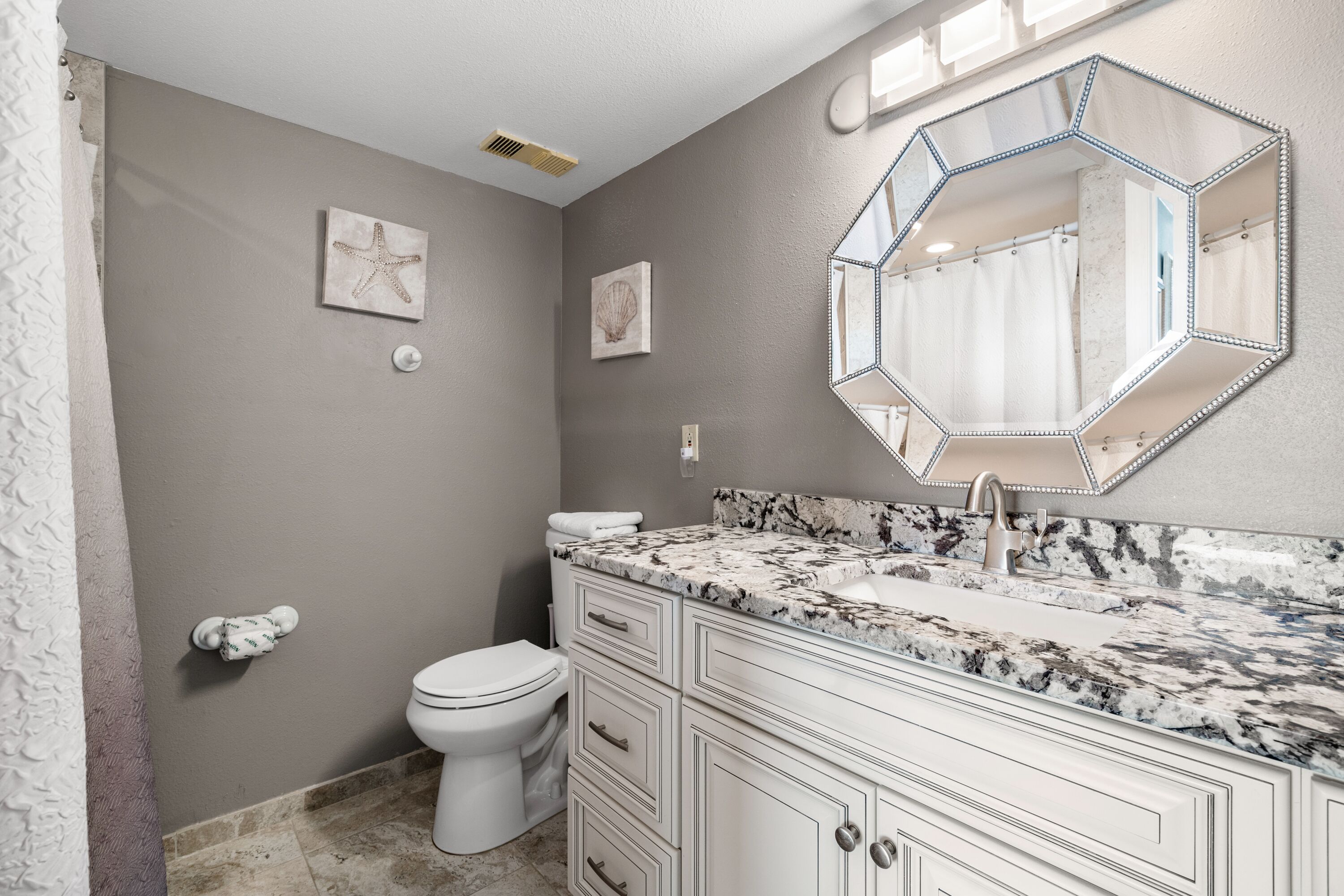 Un baño con inodoro blanco, tocador de mármol y un espejo hexagonal grande.