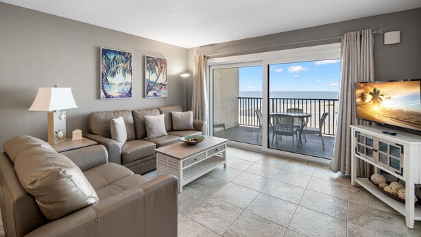 A modern living room with a sofa, coffee table, and TV, overlooking a balcony with a view of the ocean.