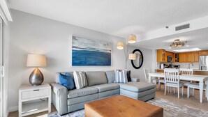Uma sala de estar moderna com um sofá seccional cinza, uma mesa de centro e um espaço de jantar com cadeiras brancas e uma mesa.