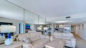 Un espacio habitable amplio con un sofá color beige, una mesa de centro y una mesa de comedor. Se ve una cocina al fondo con electrodomésticos modernos y un ventanal grande.