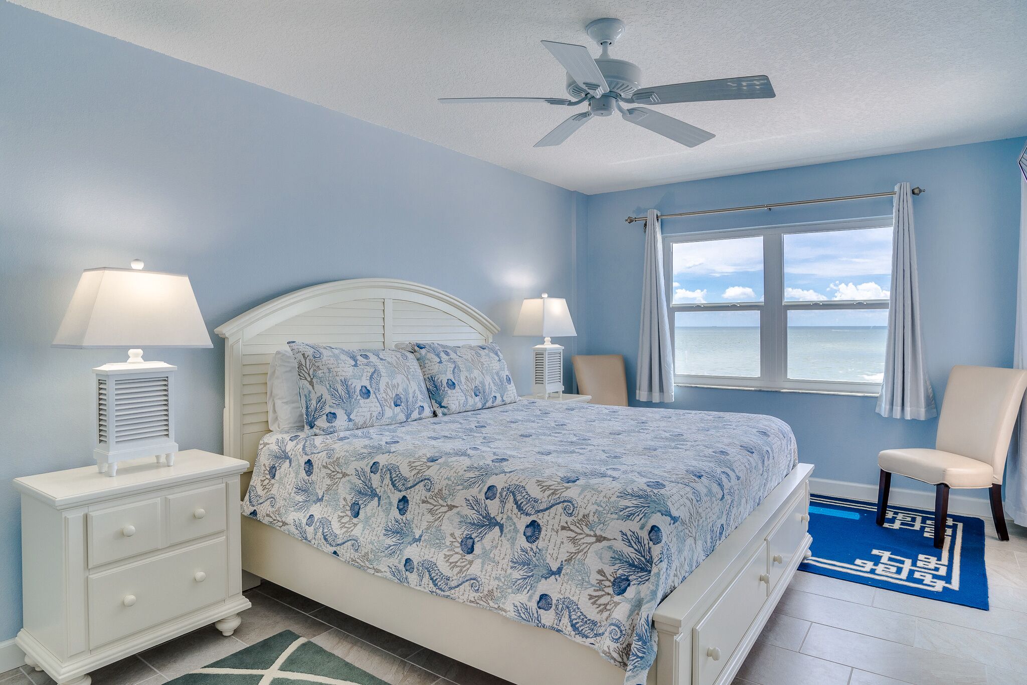 A bedroom with a bed, two bedside tables with lamps, a ceiling fan, a window with curtains, and a view of the sea.