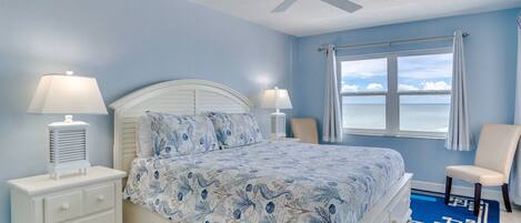 A bedroom with a bed, two bedside tables with lamps, a ceiling fan, a window with curtains, and a view of the sea.