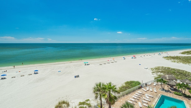 A beach with clear blue water, white sand, and a pool area with lounge chairs.