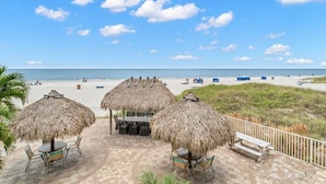 A beachside patio with thatched umbrellas and seating, overlooking a sandy beach with people and boats. - Get Away In This Well-Appointed Gulf Front Condo (Madeira Beach)