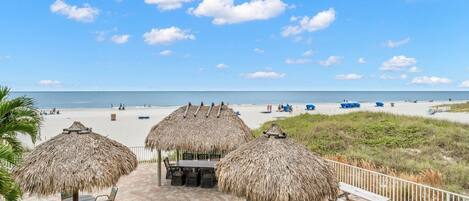 A beachside patio with thatched umbrellas and seating, overlooking a sandy beach with people and boats.