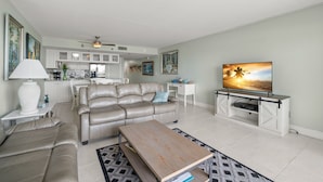 A modern living room with a sofa, coffee table, and TV stand. There is a kitchen in the background with white cabinets and a dining area. - Get Away In This Well-Appointed Gulf Front Condo (Madeira Beach)
