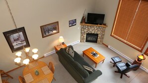 A living room with a stone fireplace, a sofa, a dining table with chairs, and a television on a stand. - Bear Creek Crossing - Your Cabin In A Condo! (Alberta)