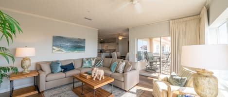 A spacious living room with a sofa, coffee table, and chairs. There is a dining area with a table and chairs visible through an open door.