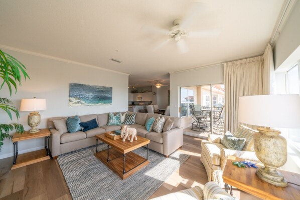 A spacious living room with a sofa, coffee table, and chairs. There is a dining area with a table and chairs visible through an open door.