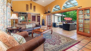 A spacious living room with a brown leather sofa, a wooden coffee table, and a patterned rug. There is a large window with a view of greenery outside. - House of Paddles, POOL, Ocean close in Keaau, Hawaii, near Hilo (Keaau)