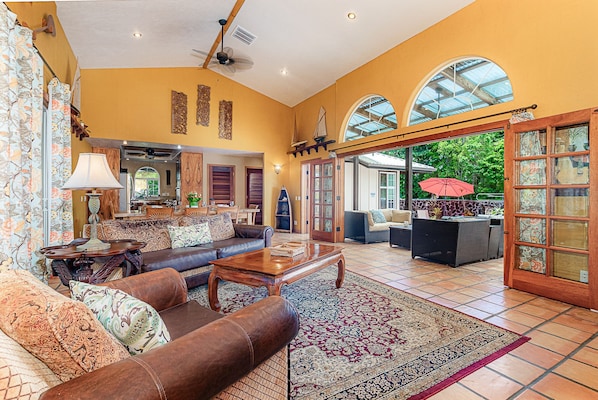 A spacious living room with a brown leather sofa, a wooden coffee table, and a patterned rug. There is a large window with a view of greenery outside.