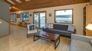Een ruime woonkamer met een houten plafond, grote ramen met uitzicht op het water, een bank, een fauteuil en een salontafel.