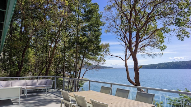 Een terras buiten met een tafel en stoelen, met uitzicht op een meer met bomen en bergen op de achtergrond.