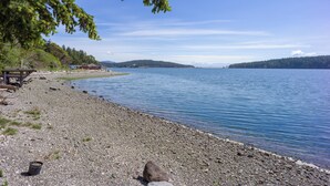Een kiezelstrand met een houten constructie en een helder uitzicht op een meer en verre bergen.