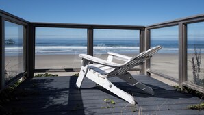 A beach chair on a wooden deck overlooking a beach with ocean waves. - Enjoy the waves in the hot tub at this beachfront home with direct beach access (Lincoln city)