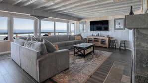 A spacious living room with a large gray sofa, a coffee table, and a patterned rug. There is a TV mounted on the wall and a stone fireplace. - Enjoy the waves in the hot tub at this beachfront home with direct beach access (Lincoln city)