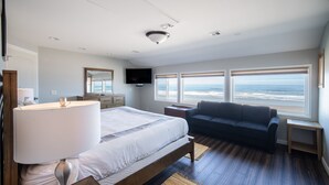 A bedroom with a large bed, a dark blue sofa, a TV, and a view of the ocean. - Enjoy the waves in the hot tub at this beachfront home with direct beach access (Lincoln city)