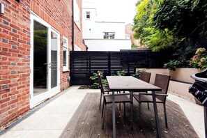 A patio with a table and chairs, a black fence, and a brick wall. - The Bohemian Rhapsody, Garden Apartment Hampstead - by Out of Office Lifestyle (London)