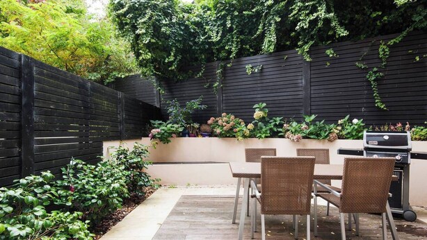A patio with a table and chairs, a grill, and a black fence with greenery.