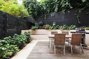 A patio with a table and chairs, a grill, and a black fence with greenery.