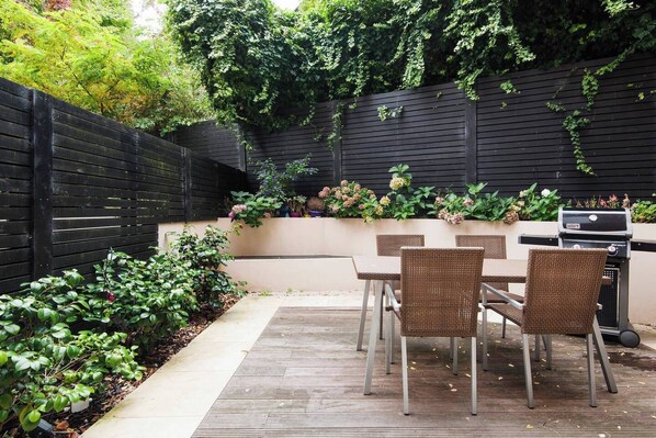 A patio with a table and chairs, a grill, and a black fence with greenery.