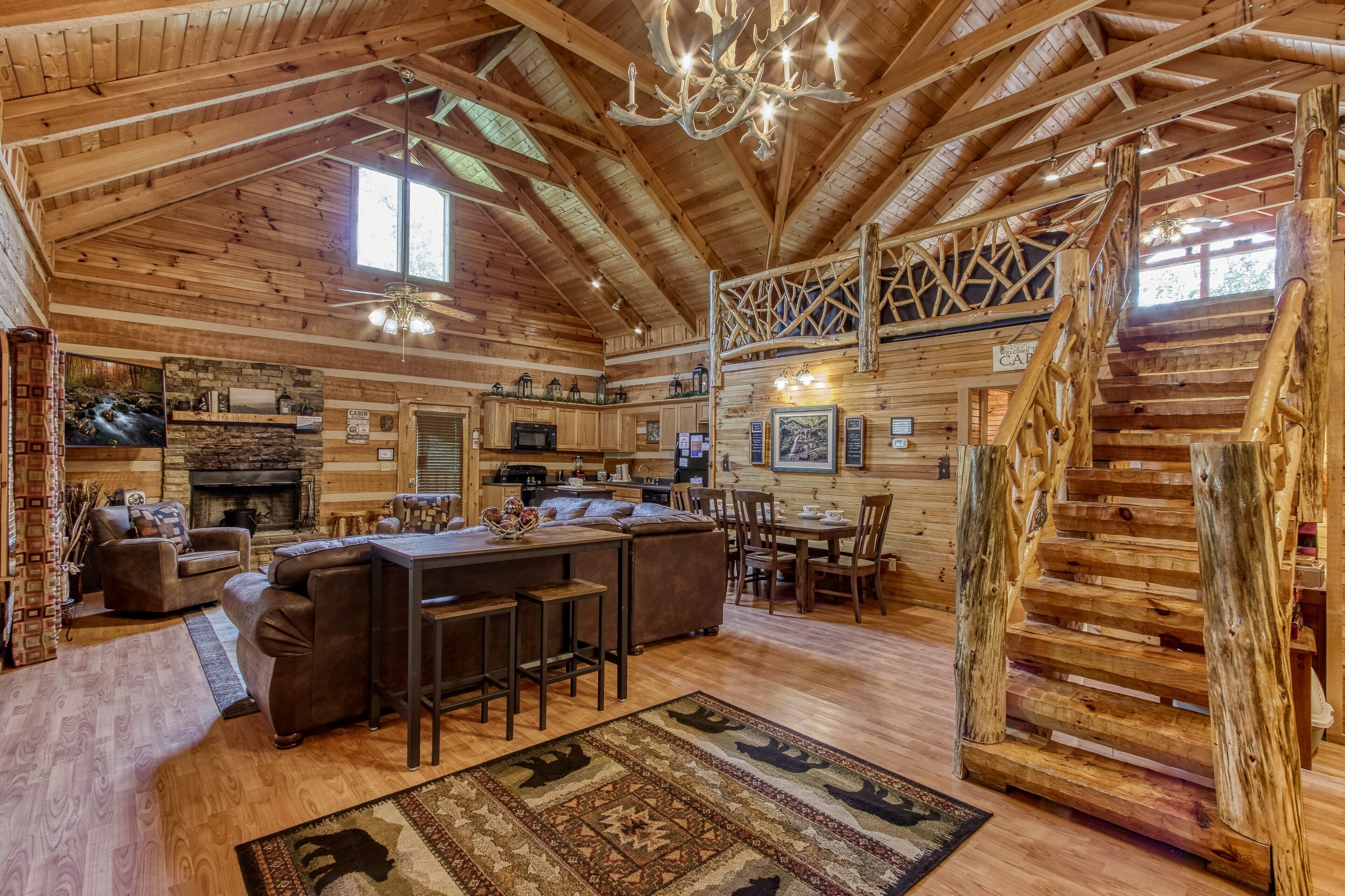 A rustic cabin interior with a wooden ceiling, a fireplace, a dining area, and a staircase.
