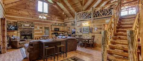 A rustic cabin interior with a wooden ceiling, a fireplace, a dining area, and a staircase.
