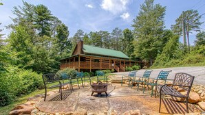 A lodge with a green roof, surrounded by a stone patio with benches and a fire pit, set in a forested area. - 4 Bedrooms Near Pigeon Forge-Hot Tub-Pet Friendly-Sleeps 12 (Sevierville)
