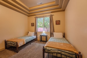 Um alojamento pequeno, de um quarto, com duas camas individuais, uma janela com cortinas, uma mesinha com abajur e dois quadros pendurados na parede.