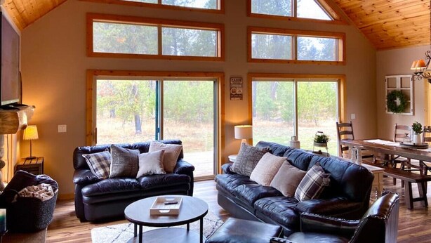 A spacious living room with a high ceiling and large windows, a fireplace, and a dining area. - Spacious Mountain Cabin w/ Hot Tub & Bunkhouse (Donnelly)