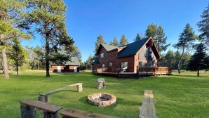A rustic cabin with a deck, surrounded by a grassy area with benches and a fire pit. - Spacious Mountain Cabin w/ Hot Tub & Bunkhouse (Donnelly)