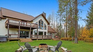 A two-story house with a deck, a fire pit with chairs, and a forested backdrop. - Luxurious Cabin with Hot Tub, Large Yard, Fire Pit (Donnelly)