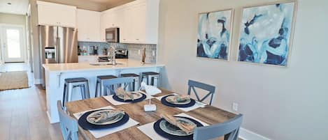 A modern kitchen with white cabinets, stainless steel appliances, and a central island. There are two abstract paintings on the wall and a dining table set with plates and napkins.