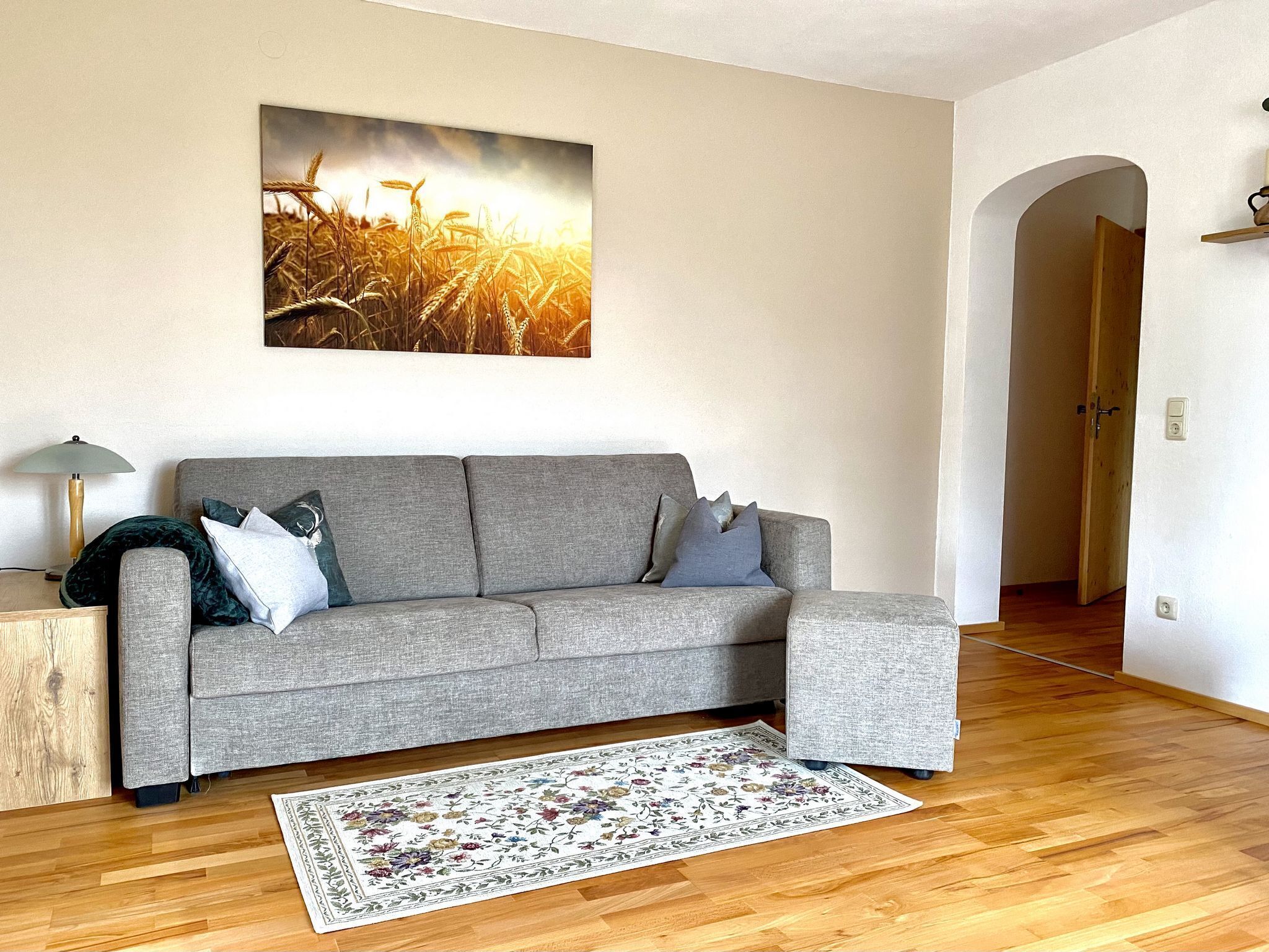 Un salon avec un canapé gris, un tapis à motifs floraux et un tableau représentant un champ de blé accroché au mur.