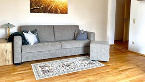 Ett vardagsrum med en grå soffa, en blommönstrad matta och en tavla som föreställer ett vetefält på väggen.