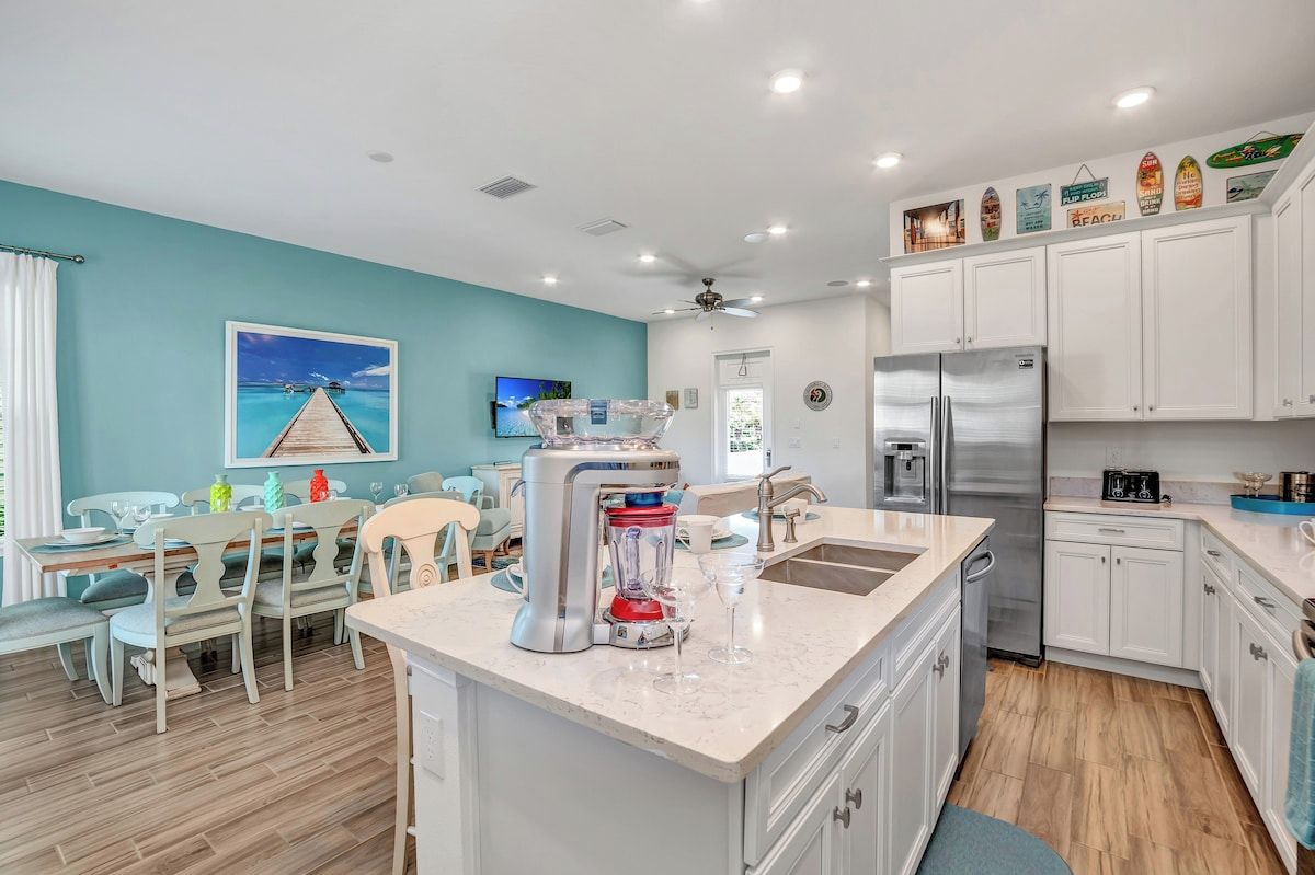 A modern kitchen with white cabinetry, a stainless steel refrigerator, a blender, and a dining area with light-colored chairs.