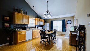 A modern kitchen with wooden cabinets, a dining area with chairs, a chandelier, and a clock on the wall.