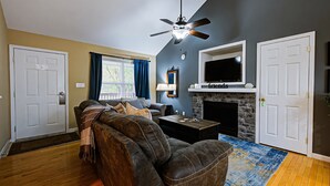 A living room with a dark brown leather sofa, a ceiling fan, a fireplace, and a television mounted above it.