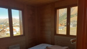 Ein Zimmer einer Holzhütte mit einem Bett, einem Heizkörper und großen Fenstern mit Blick auf eine Bergstadt.