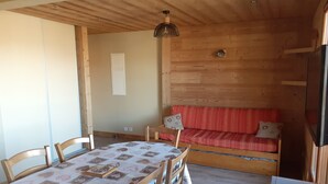 Ein gemütliches Zimmer mit Holzboden, einem Esstisch mit gemustertem Tischlauf, Holzstühlen, einer Couch mit gemusterten Kissen und einem Fernseher auf der rechten Seite.