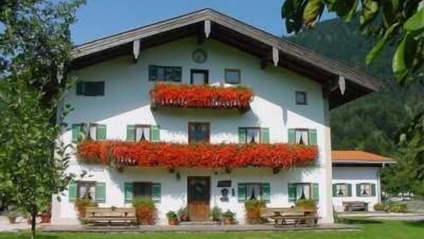 A two-story building with a balcony adorned with red flowers, a front entrance, and a small outdoor seating area. - Vacation apartment 1, up to 5 persons, 65 sqm, 2 separate bedrooms (Schleching)