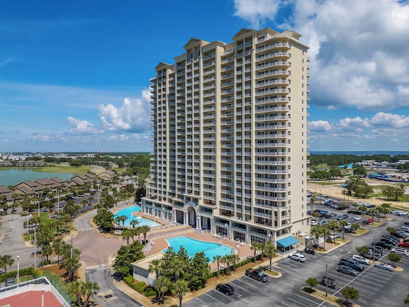 A high-rise hotel with multiple balconies, a swimming pool, and a parking lot.