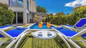 A poolside area with lounge chairs and a table, two drinks on the table, and a modern building in the background. - Villa with pool in the center of Malinska (Malinska)