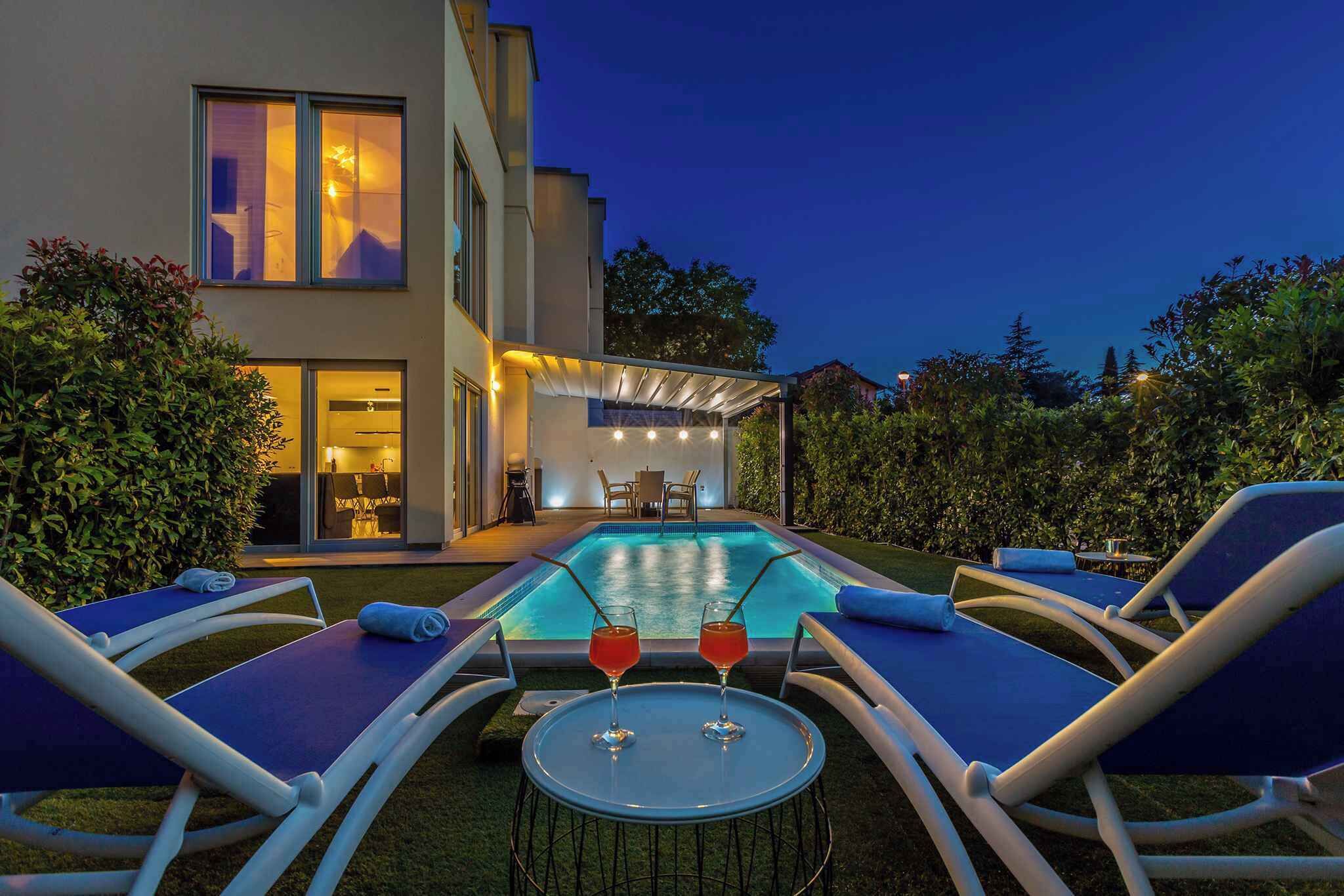A modern outdoor pool area with lounge chairs and a table with drinks, surrounded by a well-maintained garden and a building with large windows.