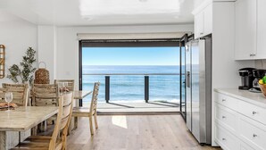 A dining area with a view of the ocean, wooden furniture, and a modern kitchen with white cabinets. - Single Family Tranquil Beachfront Rental P1829-0 (Oceanside)