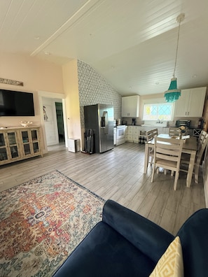 A spacious living area with a large sofa, a dining table with chairs, a kitchen with white cabinets, and a patterned rug.