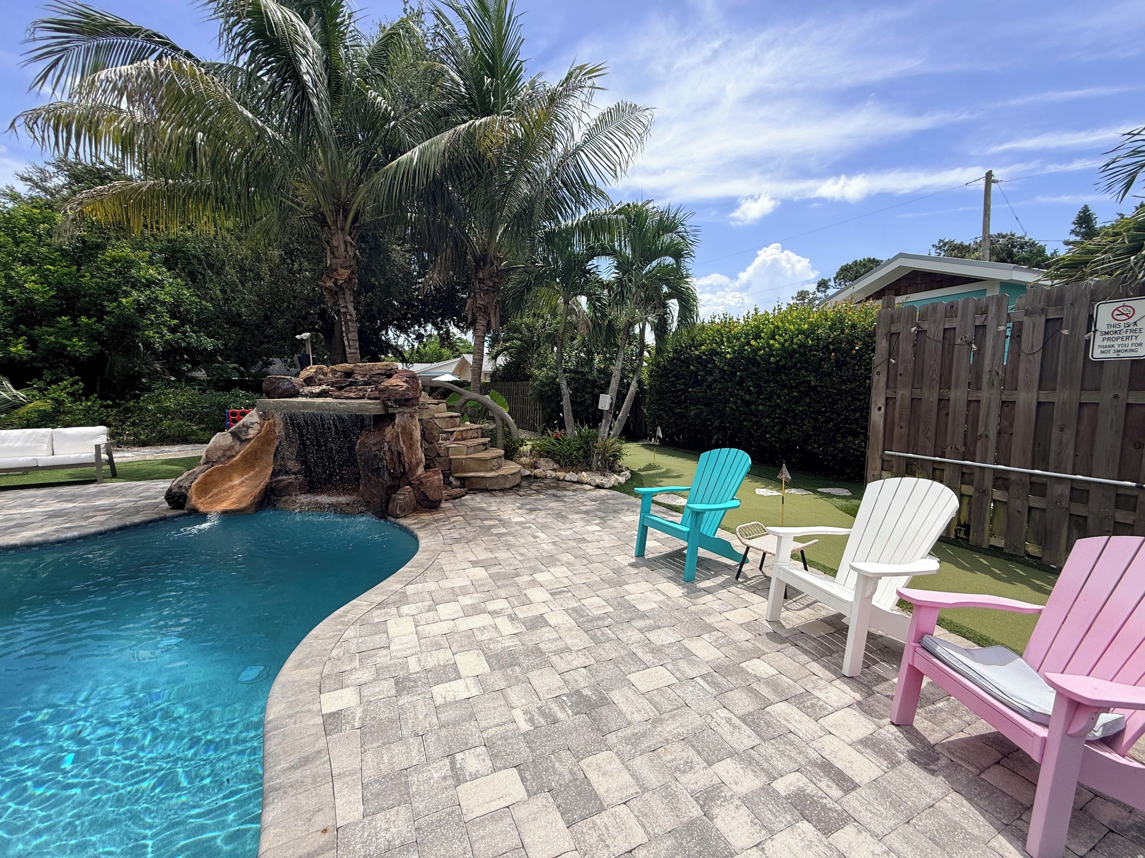 A swimming pool with a waterfall feature, surrounded by palm trees and a wooden fence.