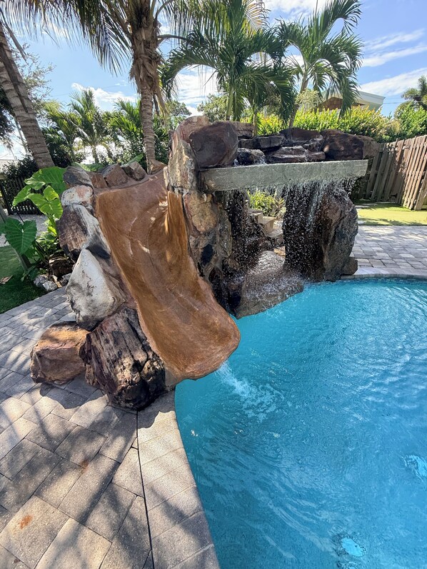A swimming pool with a rock waterfall feature and surrounding greenery.