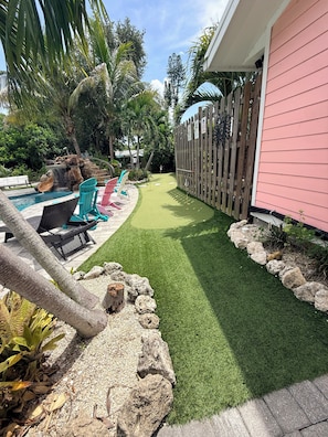 A backyard with a swimming pool, artificial grass, a wooden fence, and a pink building.
